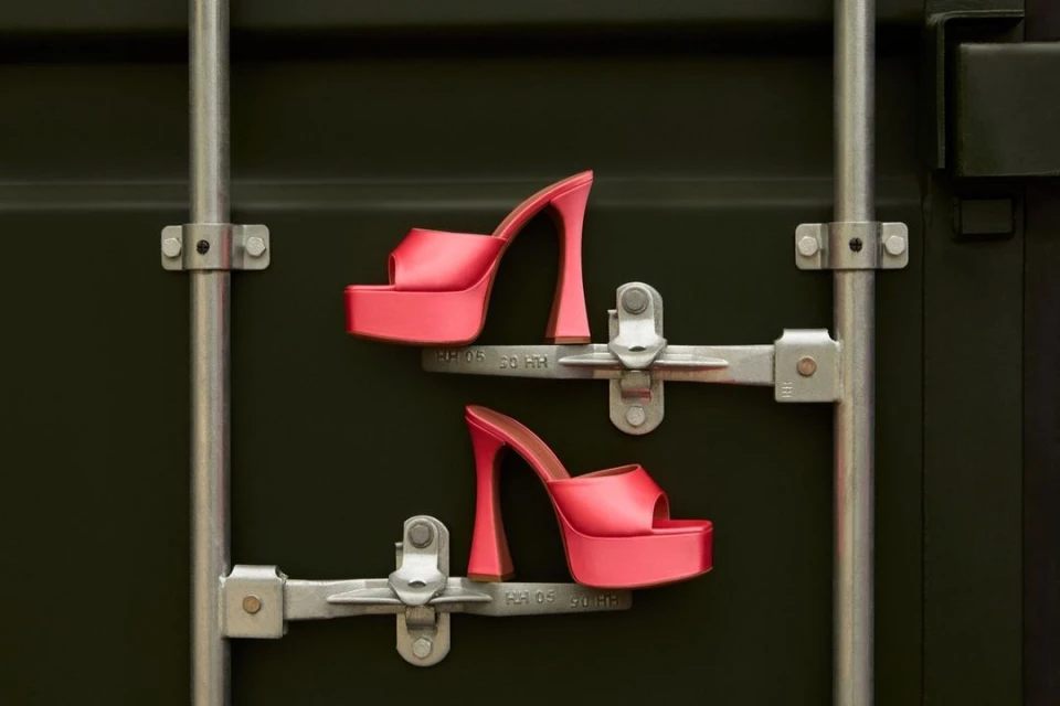 Platform high heel shoes in coral are mounted on silver metal rods against a dark green industrial container wall.