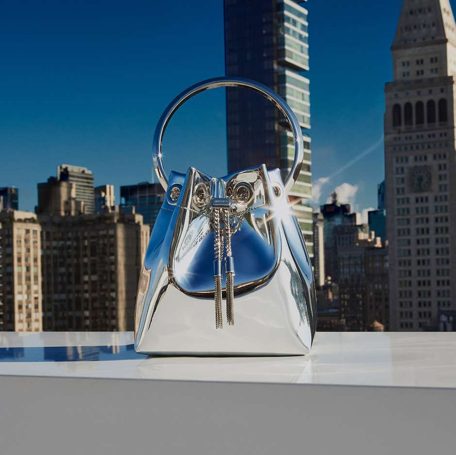 Eye-level shot of a reflective silver bucket bag with tassel detail, set against an urban skyline of tall buildings under a blue sky.