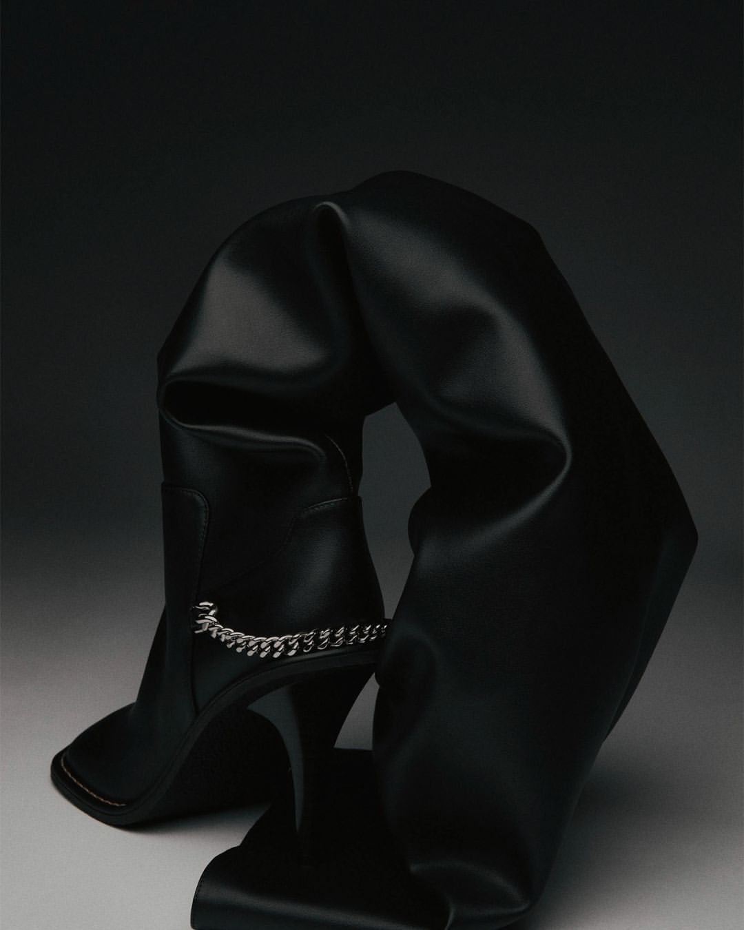 Black, over-the-knee stiletto boot is draped and folded artistically against a dark gradient background, highlighting its high heel and chain detail.