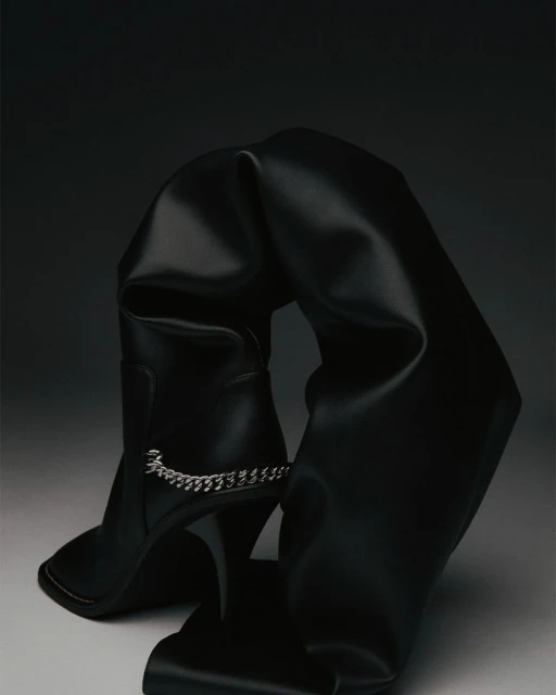 Black, over-the-knee stiletto boot is draped and folded artistically against a dark gradient background, highlighting its high heel and chain detail.