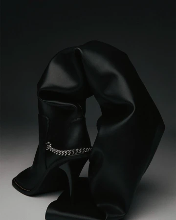Black, over-the-knee stiletto boot is draped and folded artistically against a dark gradient background, highlighting its high heel and chain detail.