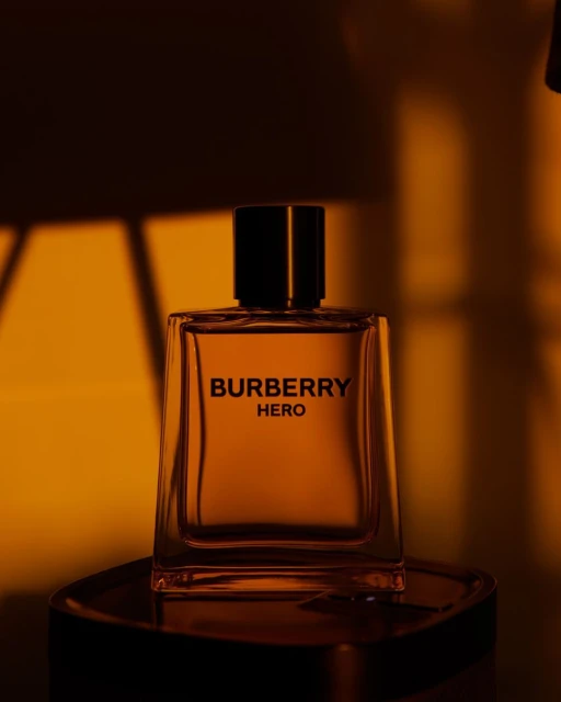 A bottle of Burberry Hero cologne sits on a dark surface against a blurred orange background, with light creating a warm, inviting atmosphere.