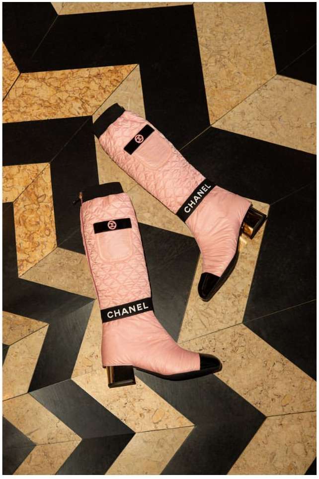 Two pale pink Chanel boots with black toe caps and logo details on a black and tan geometric tile floor in a studio setting.