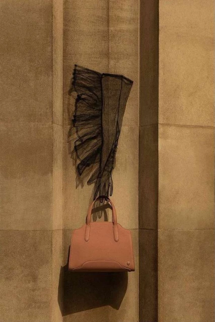 Fashion still life on a tan wall featuring a salmon-colored handbag hanging below black, ruffled, lace gloves with a soft, neutral tone.