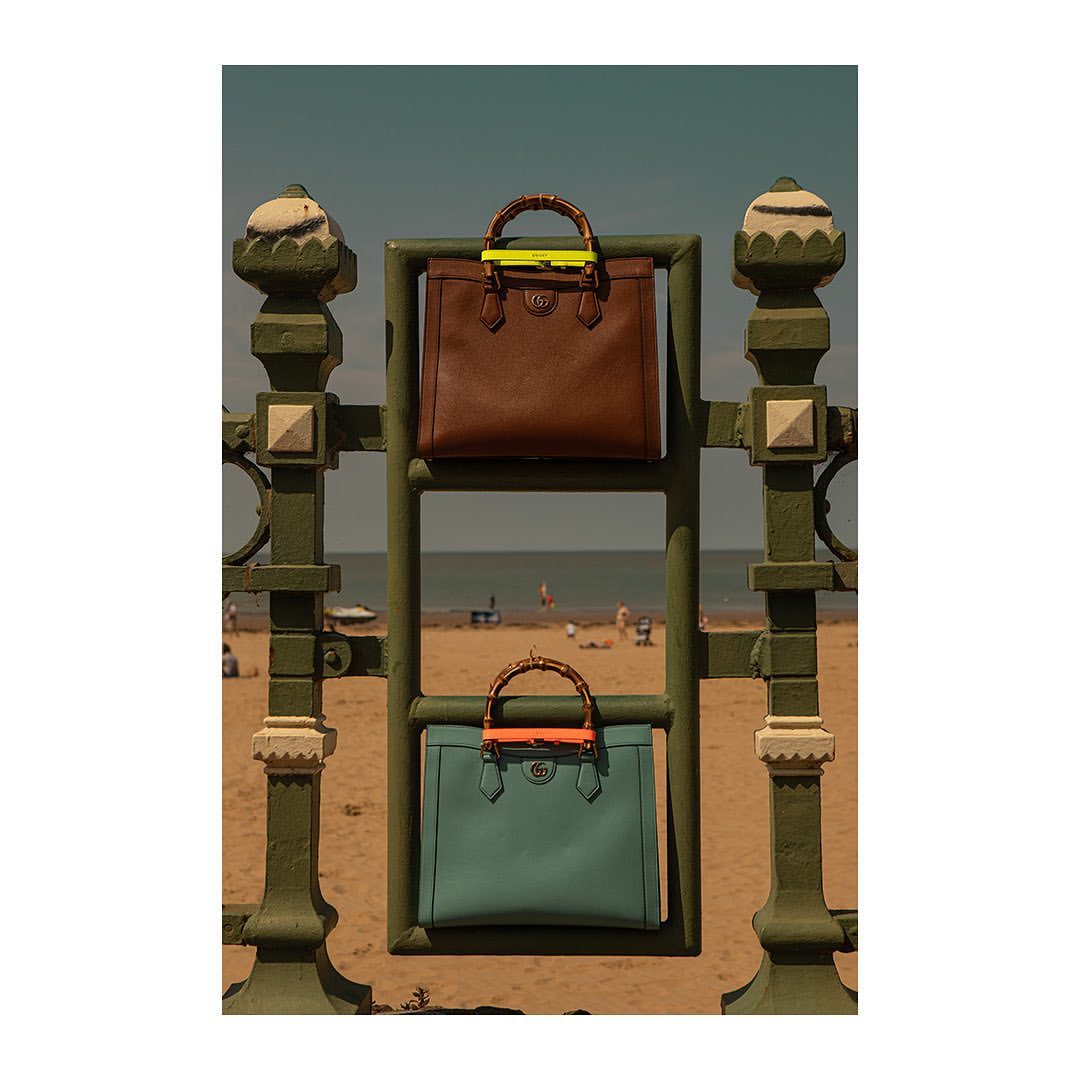 Two Gucci Bamboo tote bags, one brown, one green, are displayed on a green metal gate on a beach with the ocean and blue sky in the background.