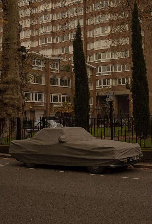 Car covered in a grey tarp parked on a street with tall trees and a large apartment building in the background on an overcast day.