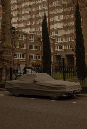 Car covered in a grey tarp parked on a street with tall trees and a large apartment building in the background on an overcast day.