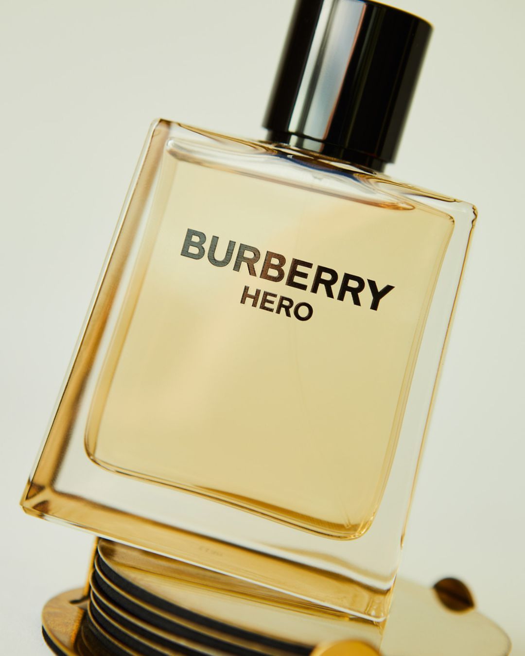 Close-up of a Burberry Hero perfume bottle. The bottle has a clear glass design with black top and gold accents, set against a light background.
