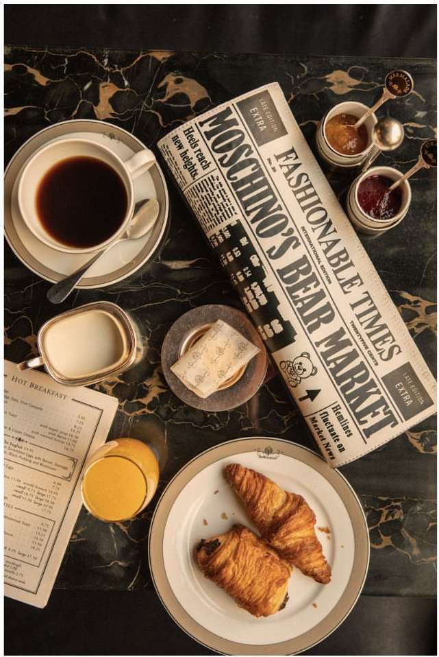 Overhead shot of a black marble table with a cup of coffee, pastries, orange juice, jam, and a newspaper featuring Moschino's Bear Market.