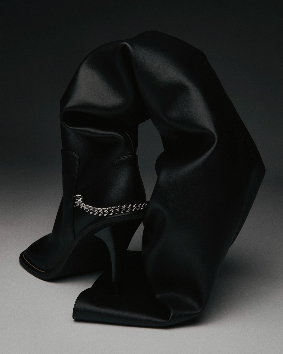 Black leather thigh-high stiletto boot with a decorative chain and folds, photographed in a studio setting with dark lighting.