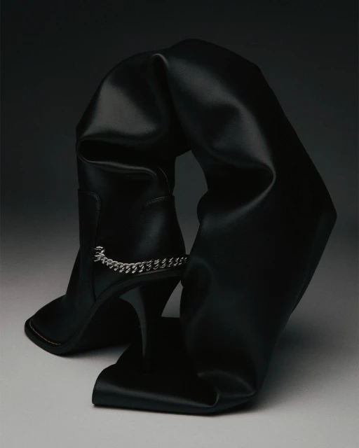 Black leather thigh-high stiletto boot with a decorative chain and folds, photographed in a studio setting with dark lighting.