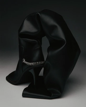 Black leather thigh-high stiletto boot with a decorative chain and folds, photographed in a studio setting with dark lighting.