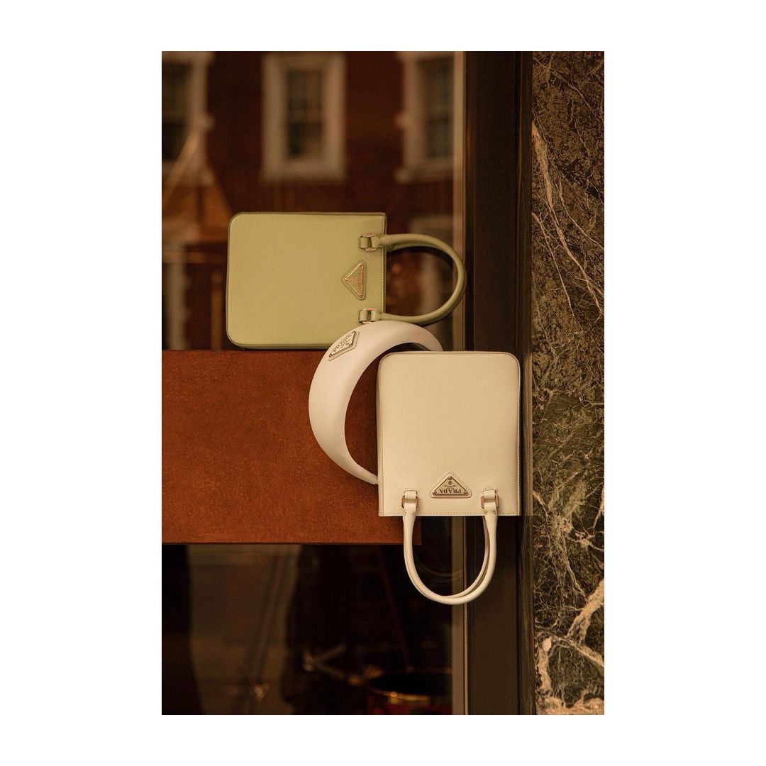 Two Prada bags, one sage and one cream, displayed in a window, showcasing their minimalist, rectangular design and elegant, curved handles.