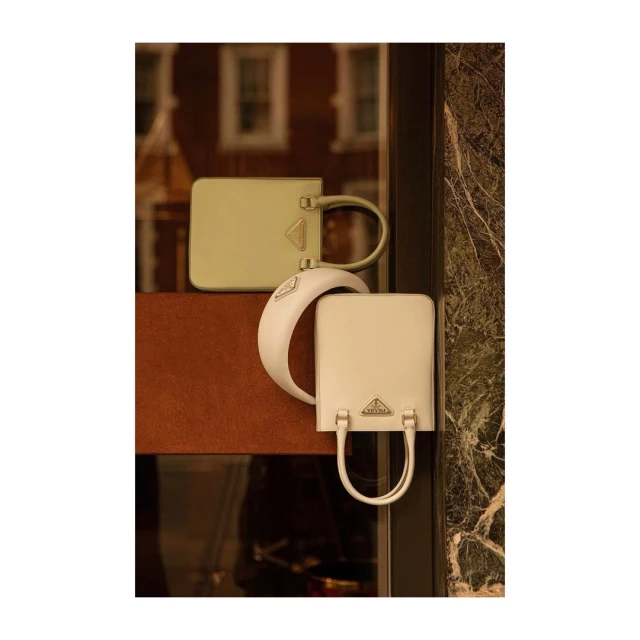 Two Prada bags, one sage and one cream, displayed in a window, showcasing their minimalist, rectangular design and elegant, curved handles.
