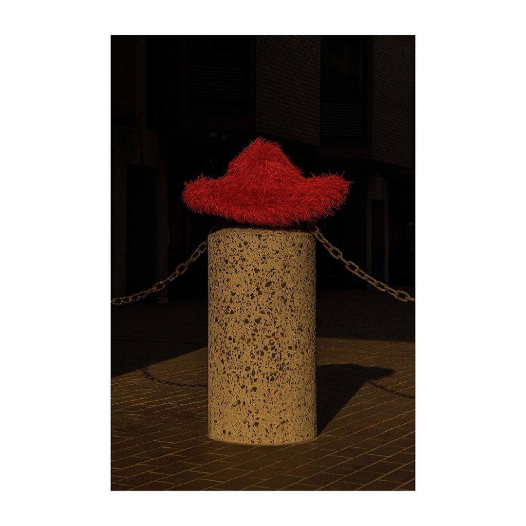 Red fuzzy hat sits atop a concrete column connected to a chain in an outdoor environment with a dark background and warm lighting.