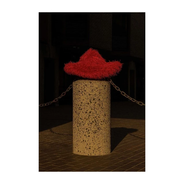 Red fuzzy hat sits atop a concrete column connected to a chain in an outdoor environment with a dark background and warm lighting.