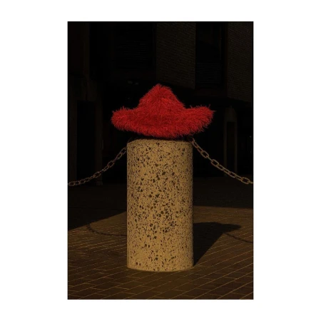 Red fuzzy hat sits atop a concrete column connected to a chain in an outdoor environment with a dark background and warm lighting.