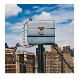 Blue leather handbag with gold hardware and a pearl strap perched against a bright blue sky and city skyline
