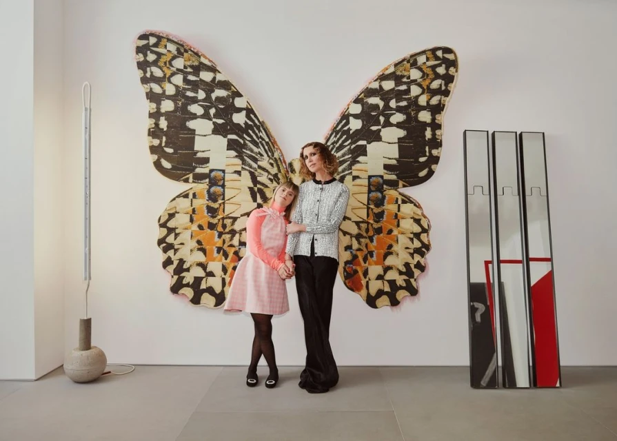 Two women pose in front of a large butterfly artwork in a white-walled studio. The older woman is in black pants and a jacket.
