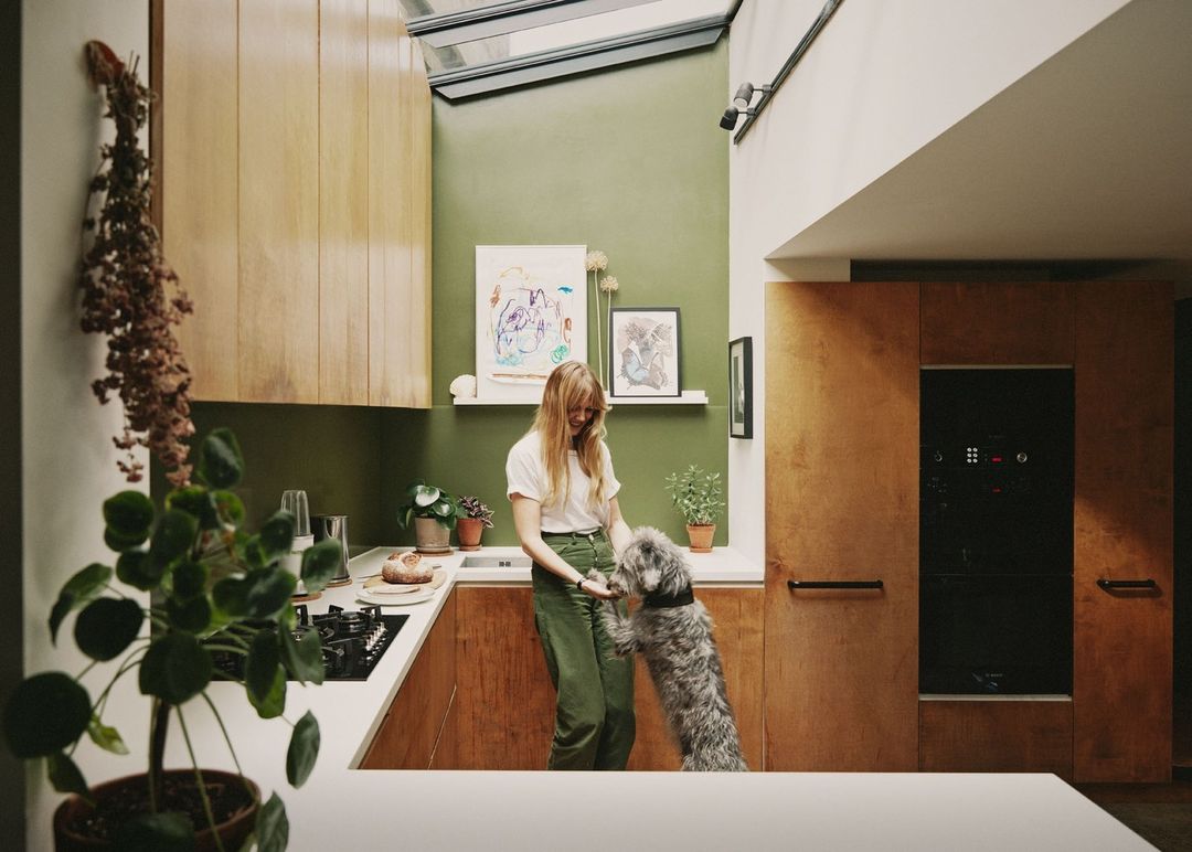 Woman interacts with her dog in a modern kitchen with pale green walls, wooden cabinets, and a skylight providing natural daylight.