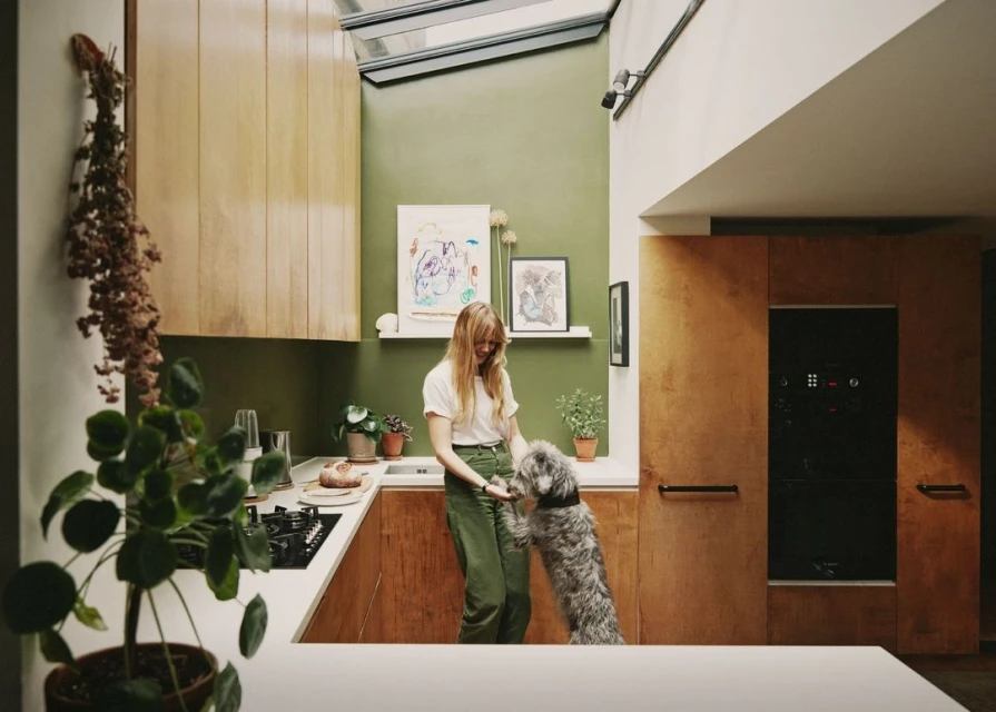 Woman interacts with her dog in a modern kitchen with pale green walls, wooden cabinets, and a skylight providing natural daylight.