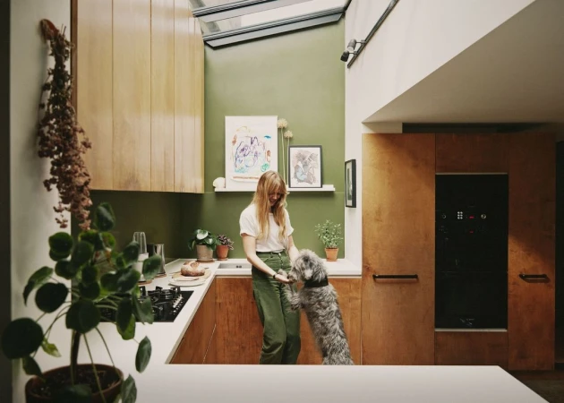 Woman interacts with her dog in a modern kitchen with pale green walls, wooden cabinets, and a skylight providing natural daylight.