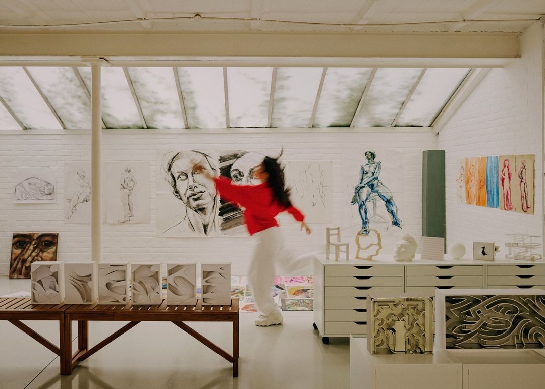 A woman in motion in an art studio, surrounded by artwork, sculptures, and light, wearing a red top and white pants.