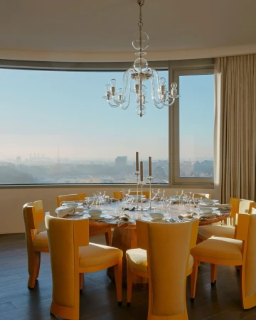 Dining room interior featuring a round wooden table set for a meal with yellow chairs, a glass chandelier, and a city view.