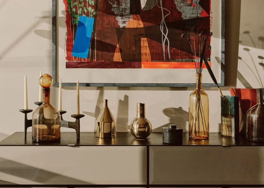 Still life features amber glass decanter, brass objects, black candlesticks, and modern art print on a gray cabinet, indoors.