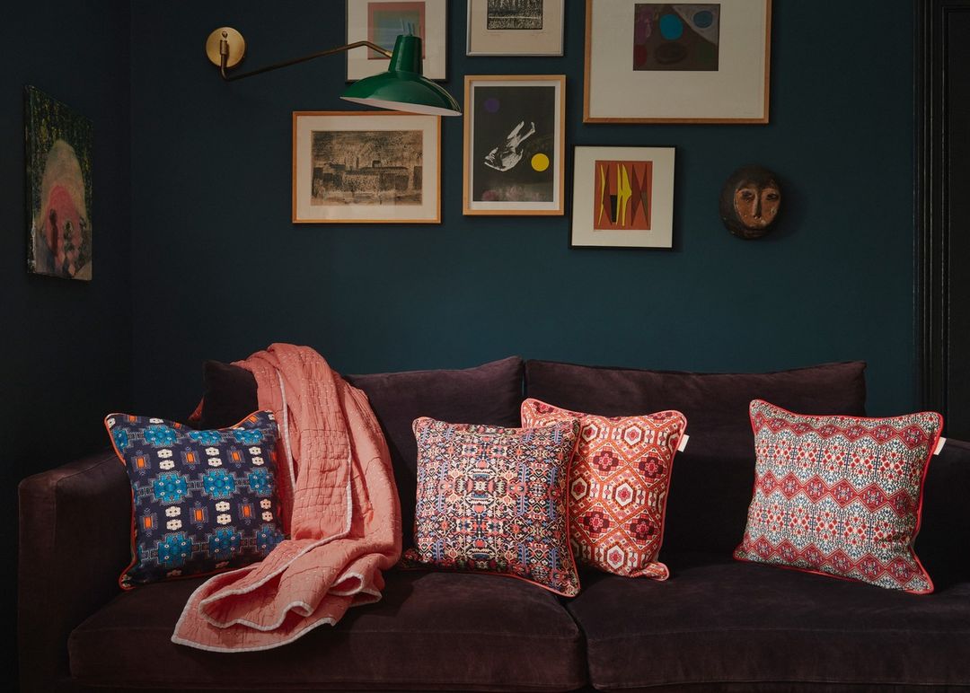Living room interior with a dark blue wall, framed art, green lamp, and a brown velvet couch adorned with patterned throw pillows.