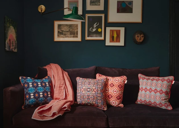 Living room interior with a dark blue wall, framed art, green lamp, and a brown velvet couch adorned with patterned throw pillows.