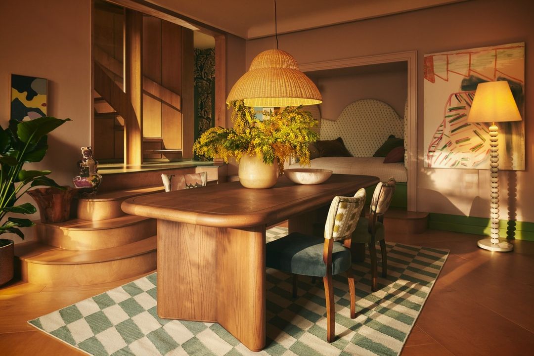Indoor dining room featuring a wooden table with a floral centerpiece, surrounded by chairs on a checkered rug, and warm lighting.