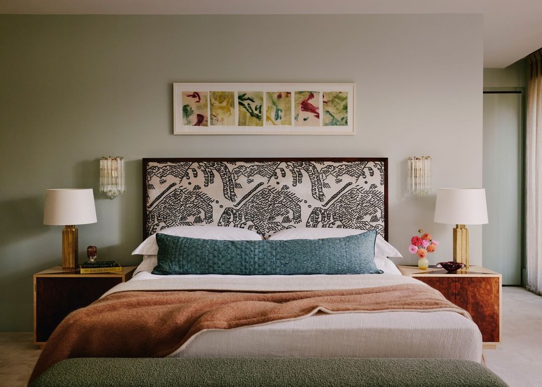 Bedroom interior with a king-sized bed, patterned headboard, and sage green walls. Nightstands flank the bed, each with a table lamp.