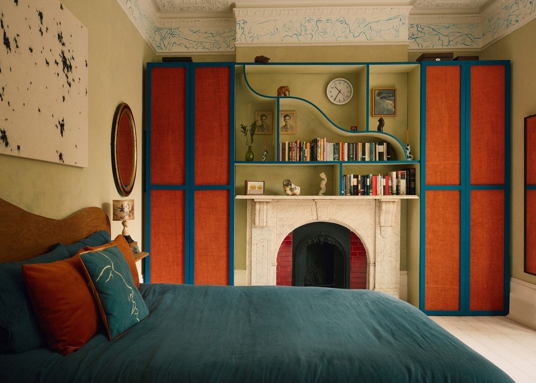 Bedroom interior featuring a bed with teal and orange pillows and duvet, complemented by a decorative mantel and colorful bookshelves.