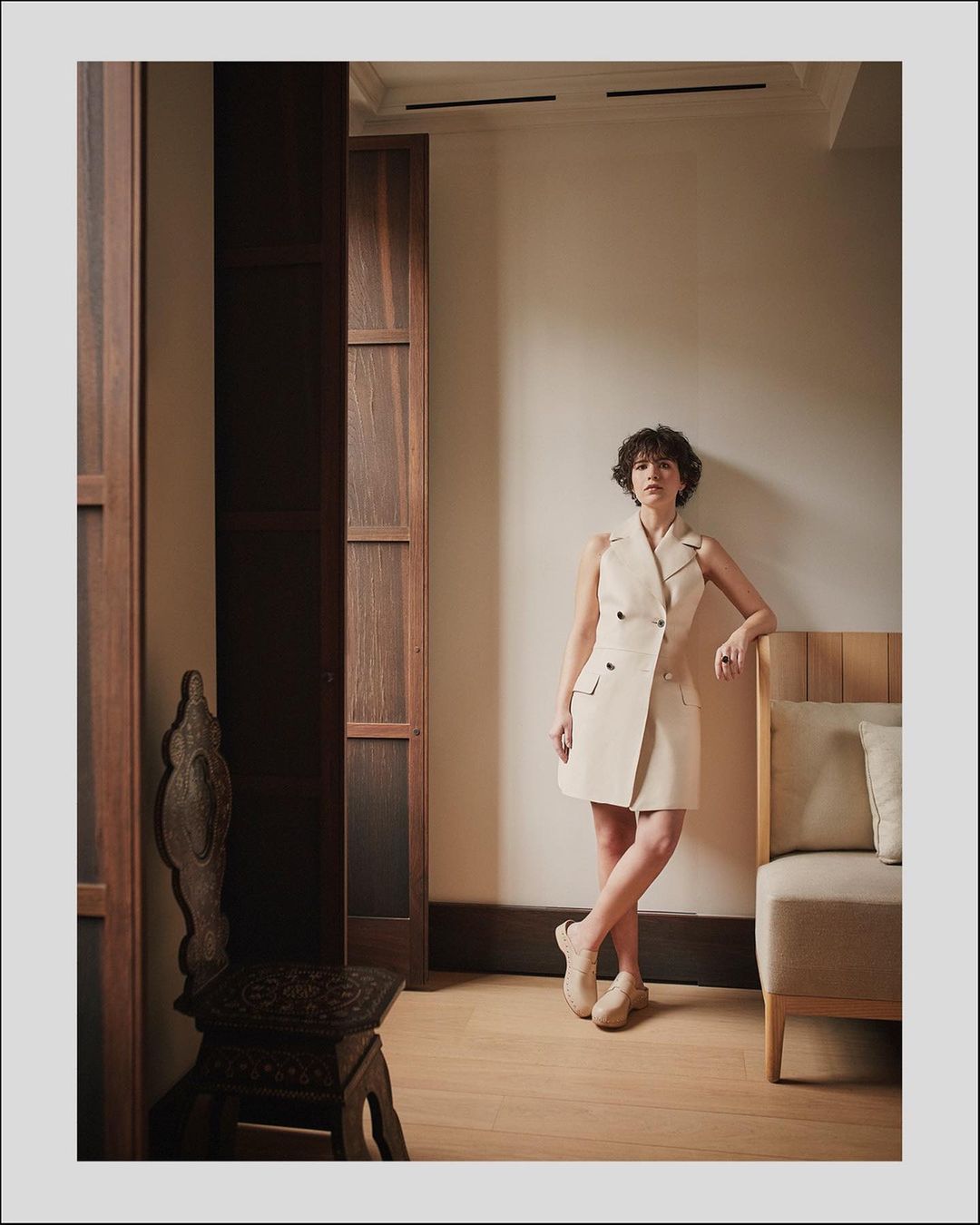 Woman in sleeveless cream vest dress leaning against an interior wall with dark wooden double doors and a modern upholstered sofa.