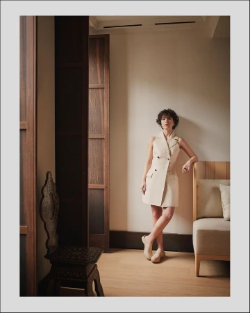 Woman in sleeveless cream vest dress leaning against an interior wall with dark wooden double doors and a modern upholstered sofa.