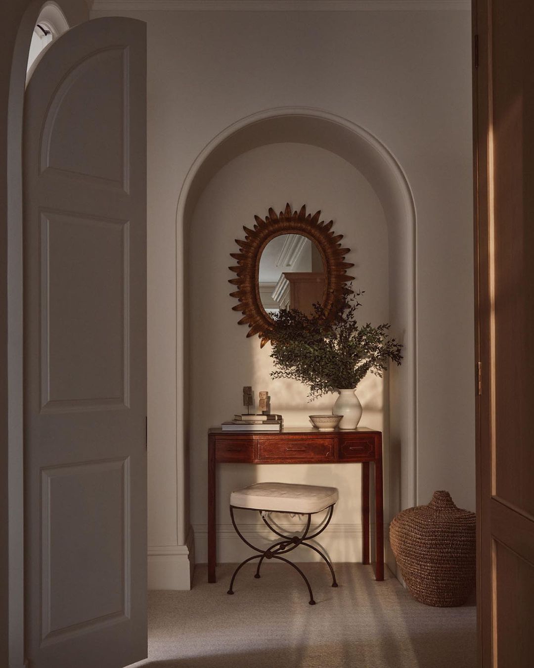 Interior view of hallway with console table, stool, mirror, vase with green branches, and decorative basket in soft natural light.