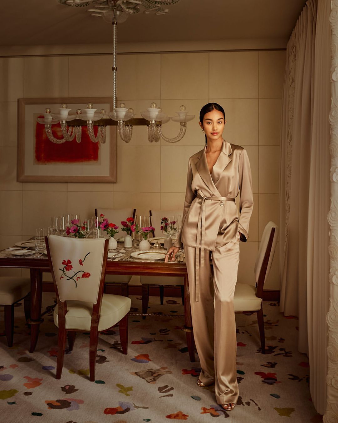 A slender woman in a bronze silk suit stands in a dining room with a table set for dinner, soft light and elegant floral carpet.