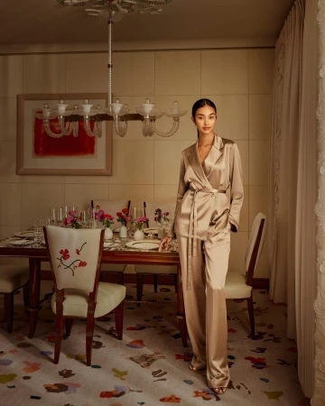 A slender woman in a bronze silk suit stands in a dining room with a table set for dinner, soft light and elegant floral carpet.