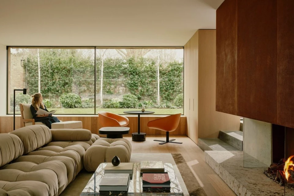 Living room with a beige sofa, orange chairs, and a fireplace, woman reading by window, showcasing cozy modern interior design with a garden view.
