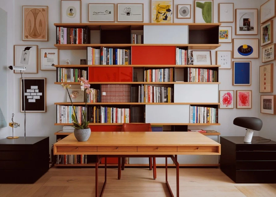 Home office interior with a light wood desk, a wall of bookshelves filled with books, and framed art in a brightly lit room.