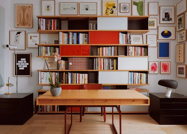 Home office interior with a light wood desk, a wall of bookshelves filled with books, and framed art in a brightly lit room.