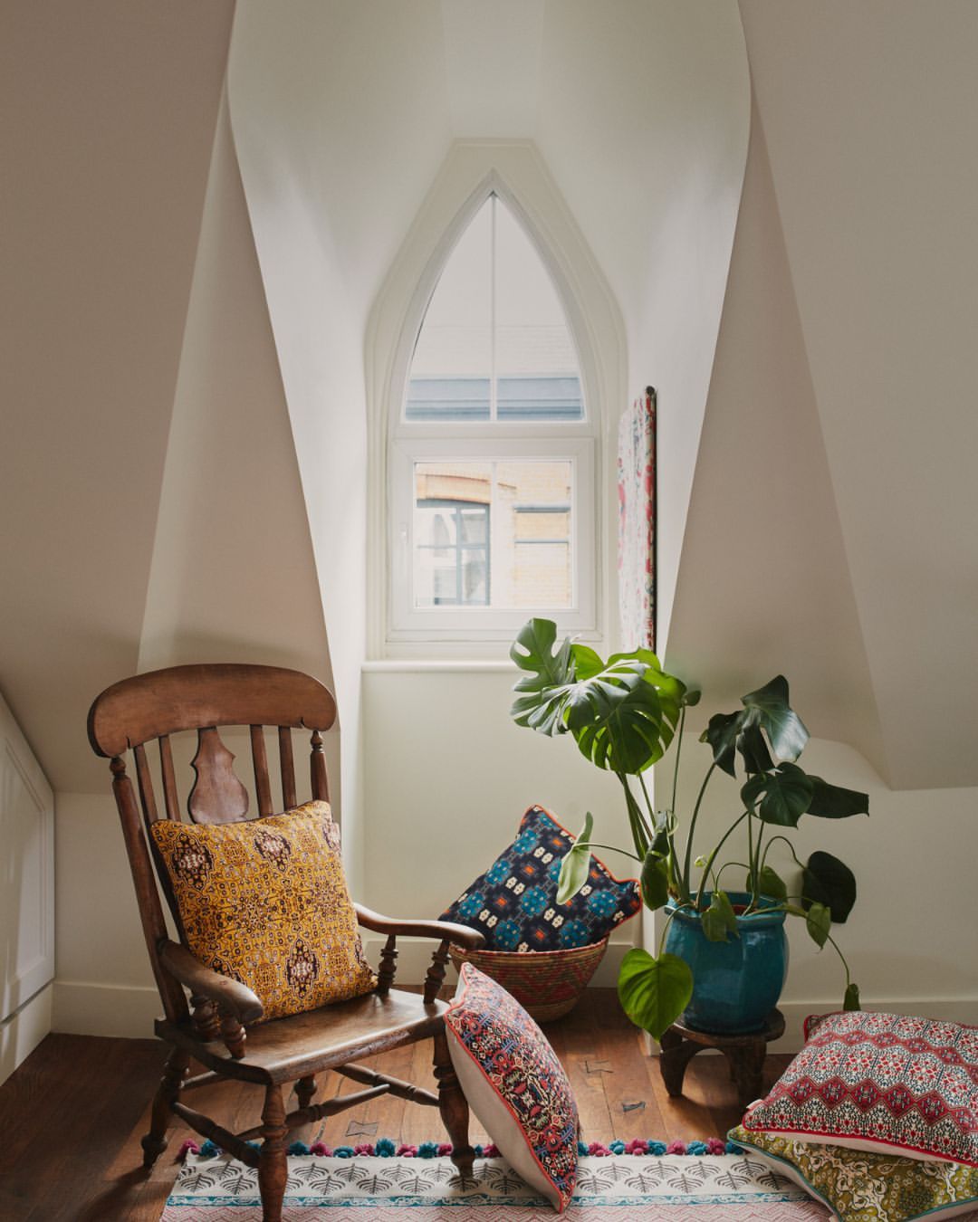 Cozy interior with wooden chair, patterned pillows, and a potted monstera plant near a window in an attic room with sloped ceilings.