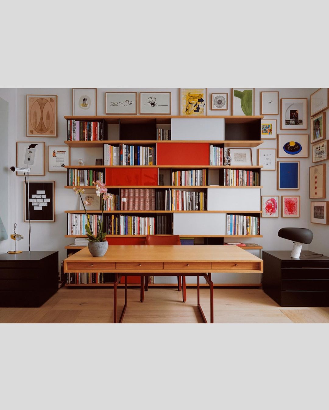 Interior view of a home office with a wooden desk, a bookshelf filled with books and decorative objects, and various framed artworks on the wall.