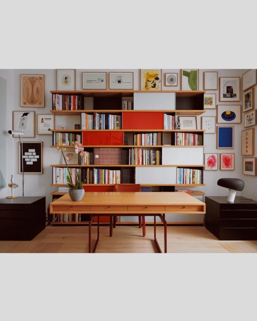 Interior view of a home office with a wooden desk, a bookshelf filled with books and decorative objects, and various framed artworks on the wall.