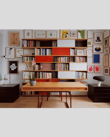 Interior view of a home office with a wooden desk, a bookshelf filled with books and decorative objects, and various framed artworks on the wall.