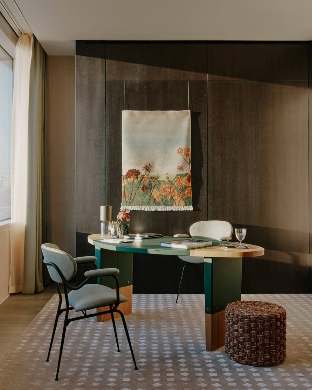 A modern home office features a green and wood desk with flowers, books, and a water glass, complemented by a gray chair and stool.