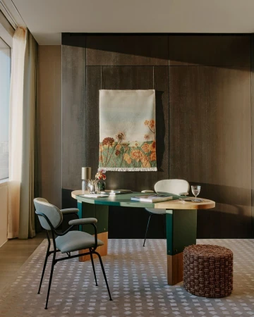 A modern home office features a green and wood desk with flowers, books, and a water glass, complemented by a gray chair and stool.