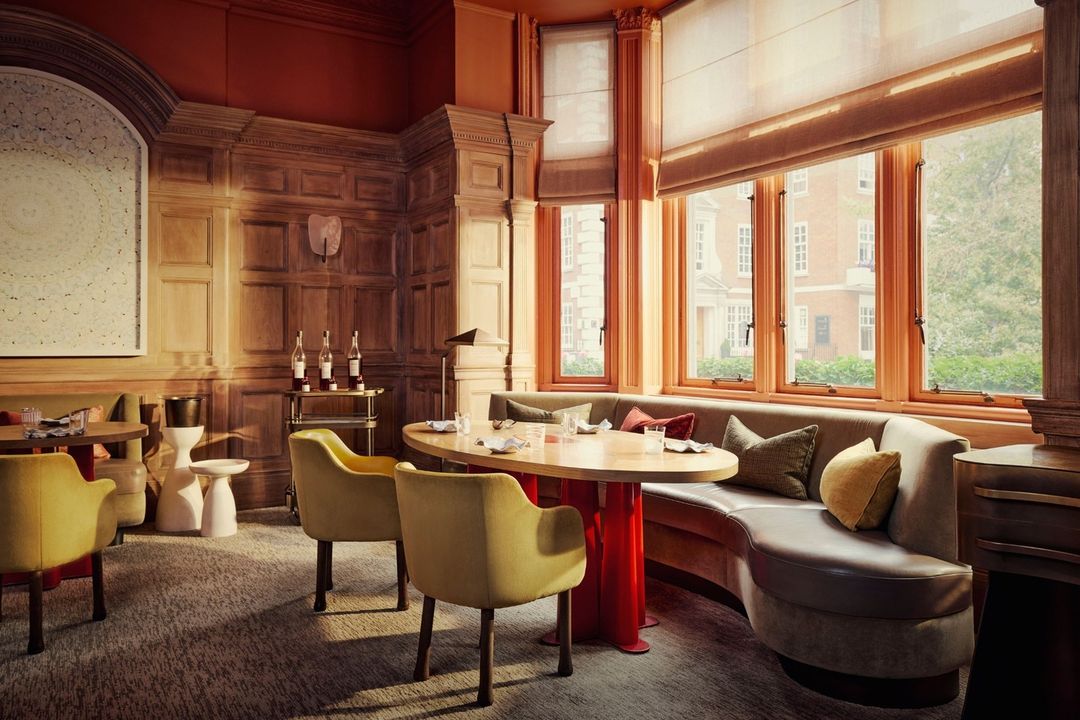 Interior of a warmly lit restaurant with ochre chairs around a round table, set against wood-paneled walls and large windows.