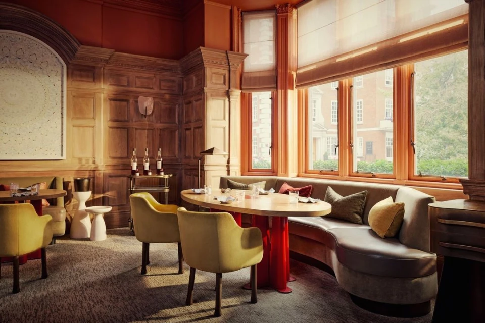 Interior of a warmly lit restaurant with ochre chairs around a round table, set against wood-paneled walls and large windows.